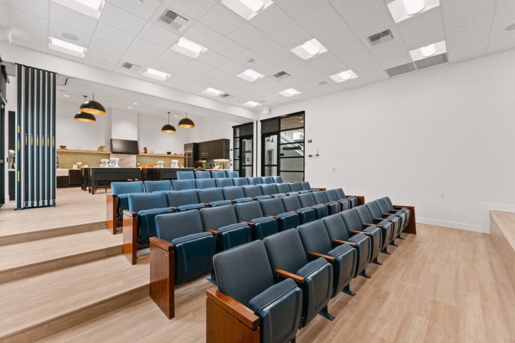 Community Lecture Room with tiered blue seating, wooden armrests, and an adjacent open kitchen/lounge area in a modern apartment complex.